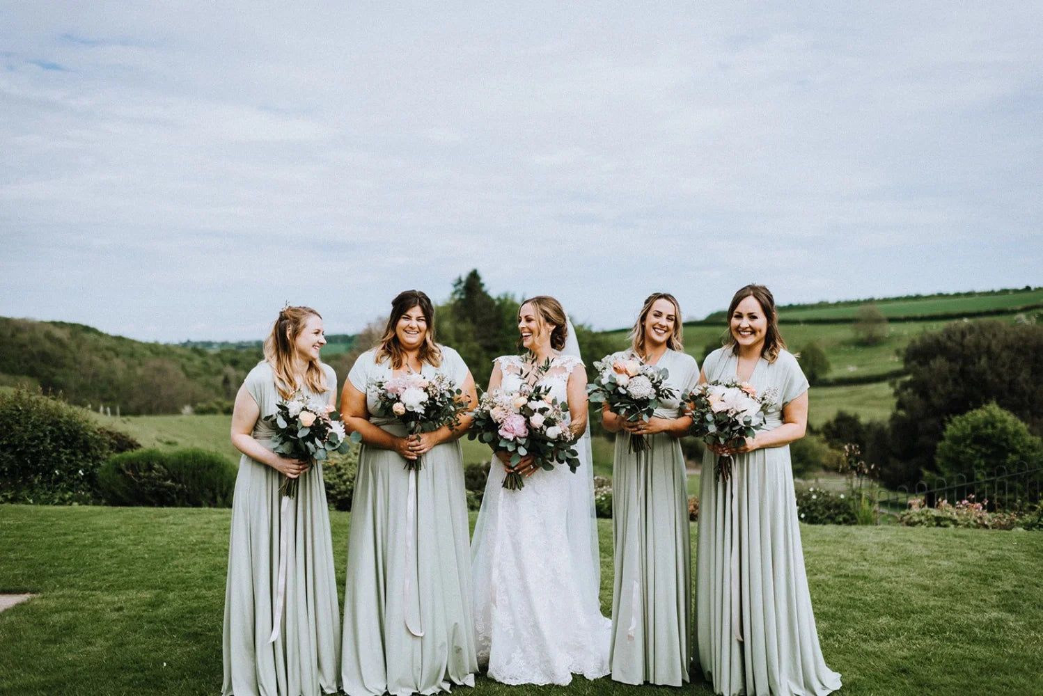 Five women in formal green bridesmaid infinity gowns holding bouquets of flowers