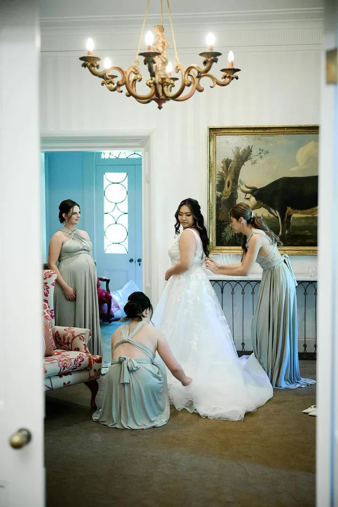 Bride in a white infinity gown assisted by bridesmaids in sage green convertible dresses