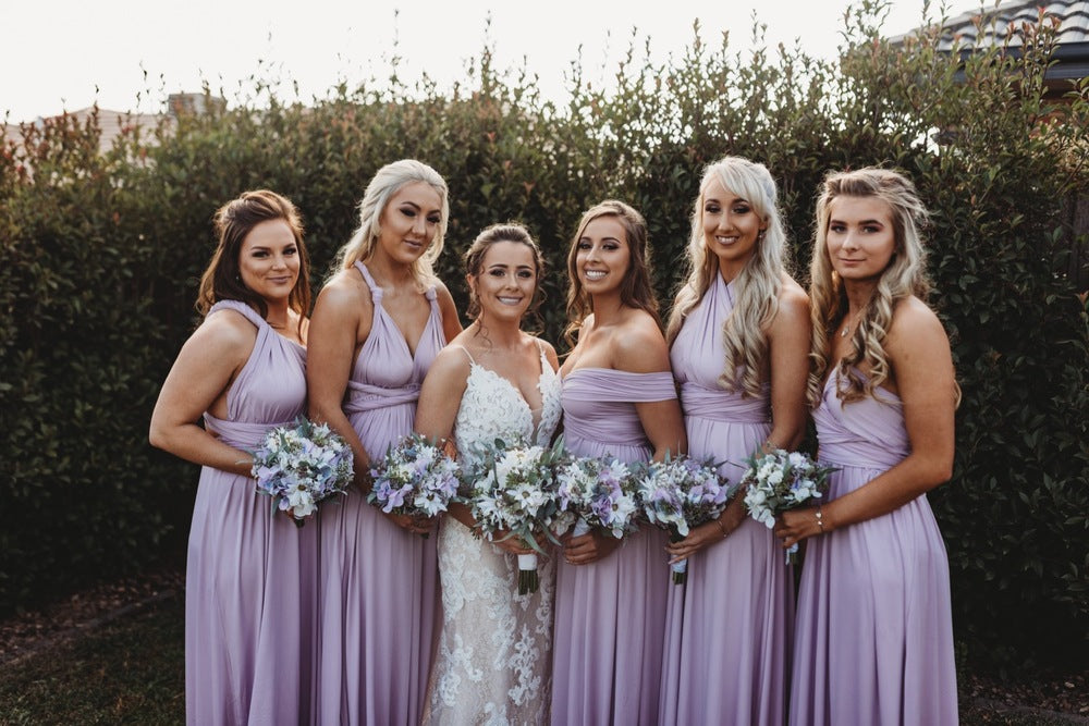 Six women in lavender bridesmaid dresses and a white wedding gown holding bouquets, showcasing the Lilac Infinity Gown and bridesmaid dresses