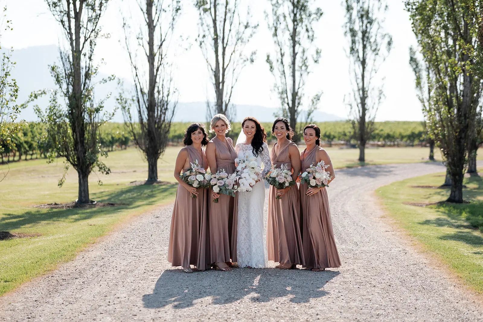 Bride in white lace gown with five taupe infinity bridesmaid dresses holding floral bouquets