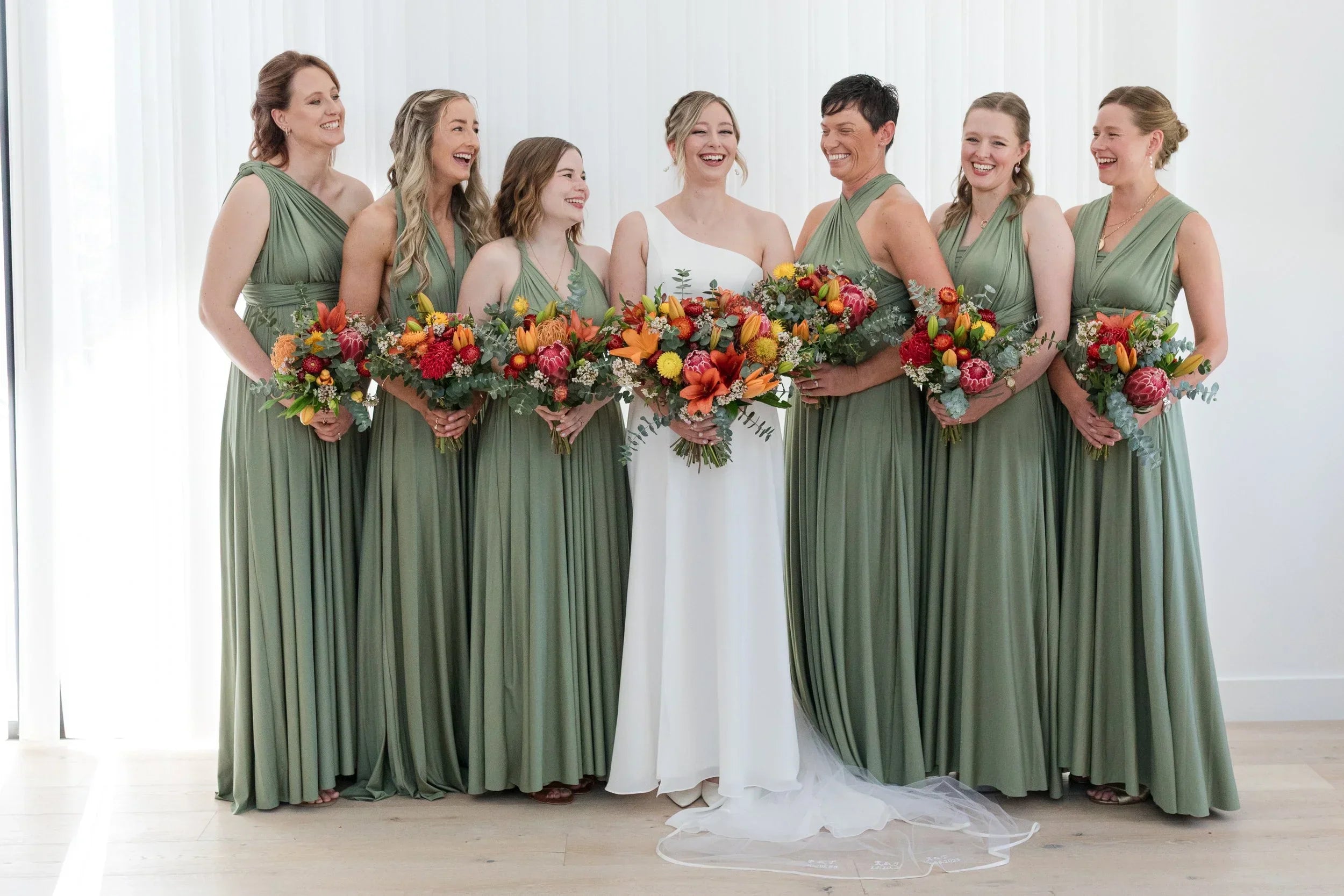 Seven women in wedding party with bride in white and bridesmaids in sage green infinity bridesmaid dresses holding colorful bouquets
