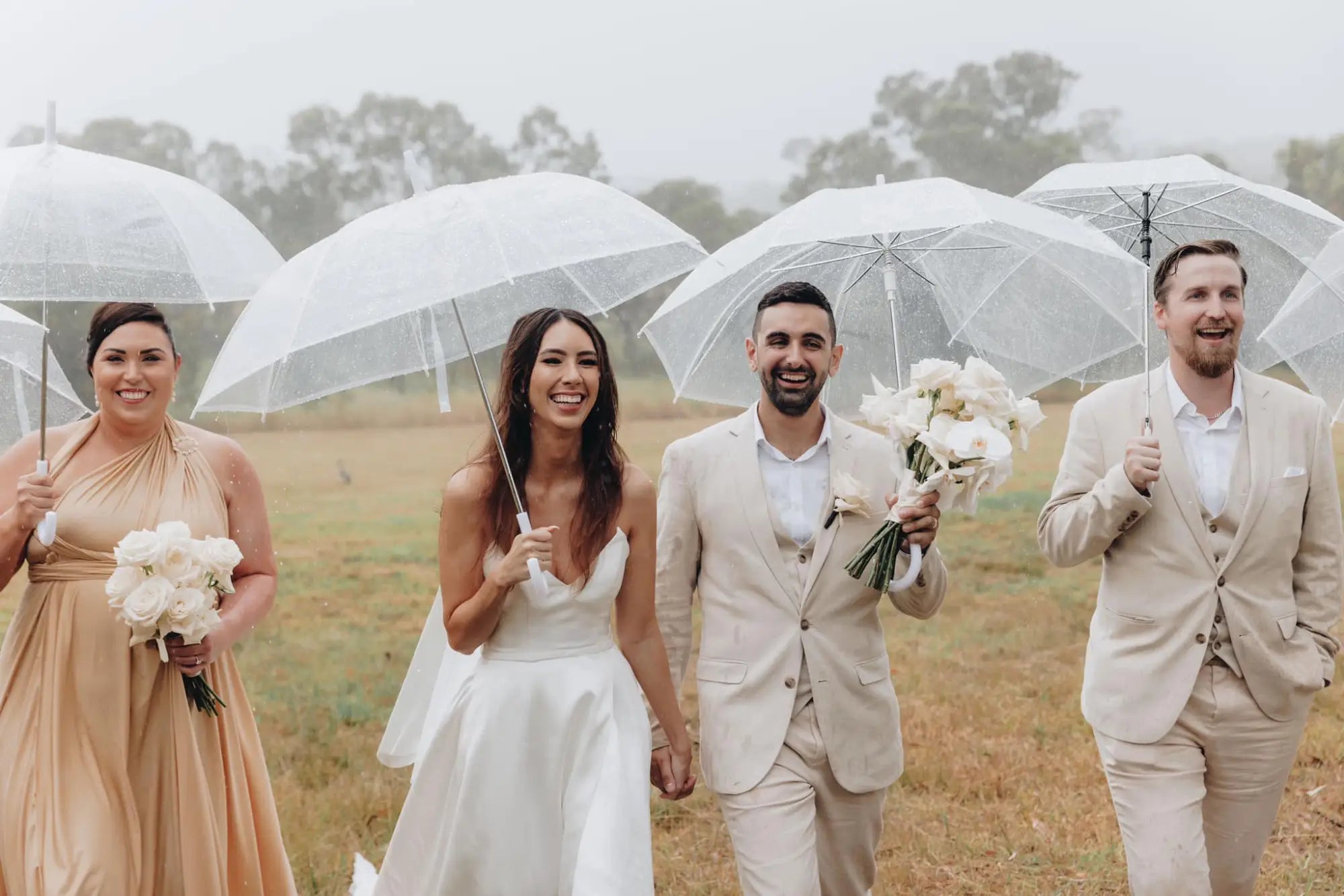Wedding party walking outdoors in rain with clear umbrellas, showcasing infinity bridesmaid dress