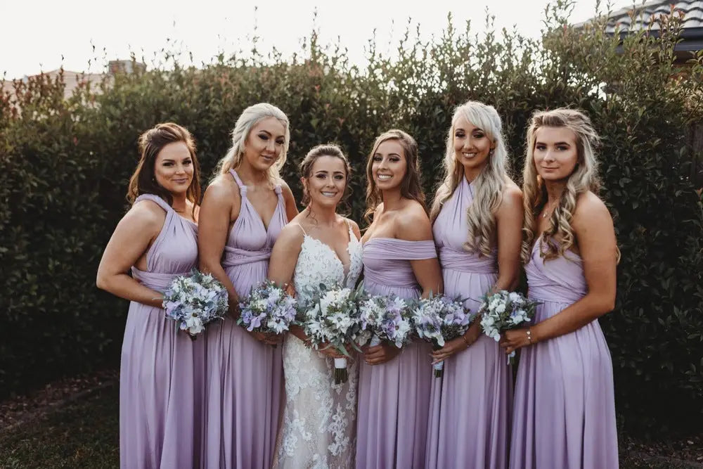 Six women in lavender infinity dresses and a bride in a white lace gown with bouquets