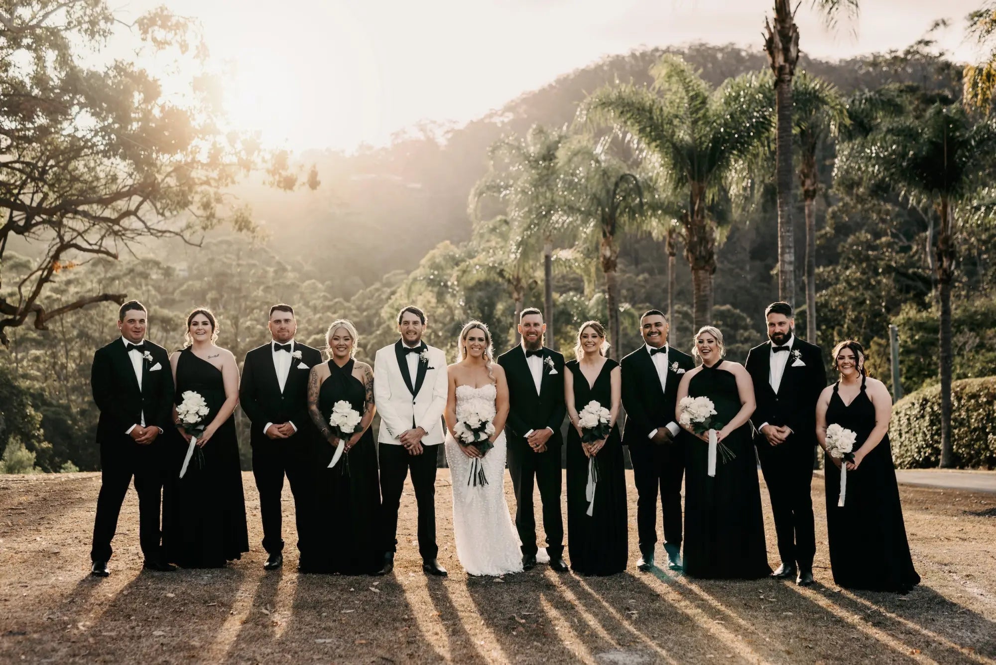 Wedding party with bride in white strapless gown and groom in white tuxedo, bridesmaids in black dresses, groomsmen in tuxedos, Luxe Satin Black