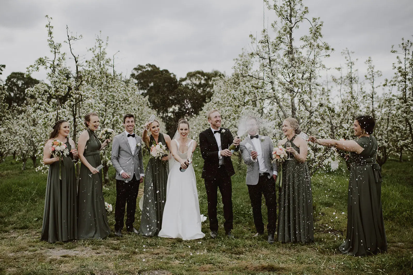Wedding party celebrating in an orchard wearing Luxe Satin Olive infinity bridesmaid dress