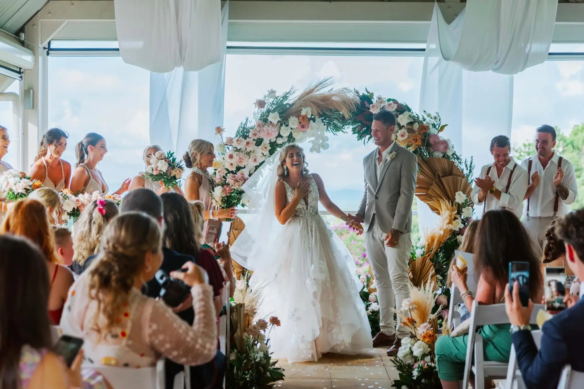 Joyful bride and groom holding hands under floral arch at Olivia and Reece wedding with infinity dress