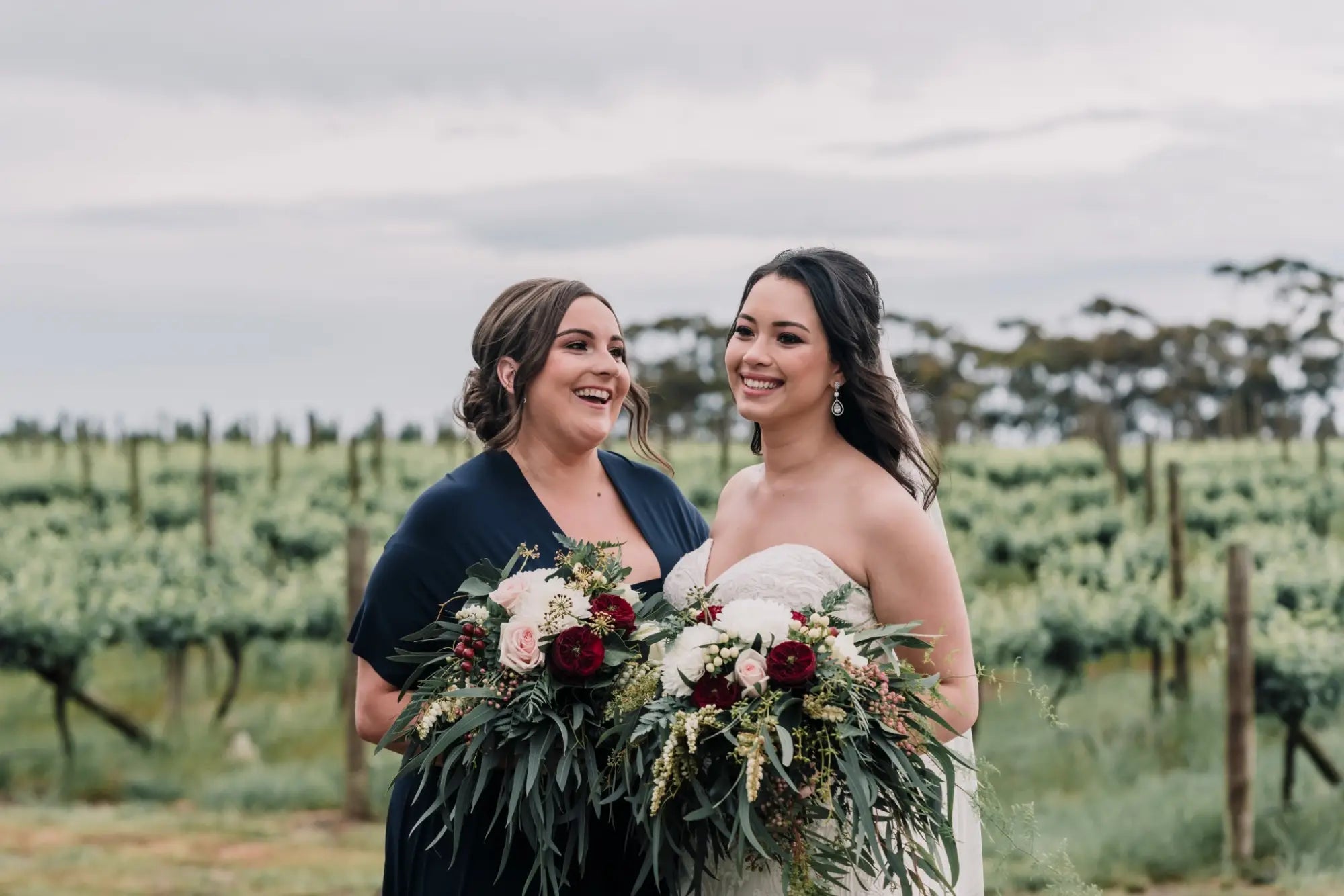Two smiling women in wedding attire holding bouquets in a vineyard, wearing infinity dresses