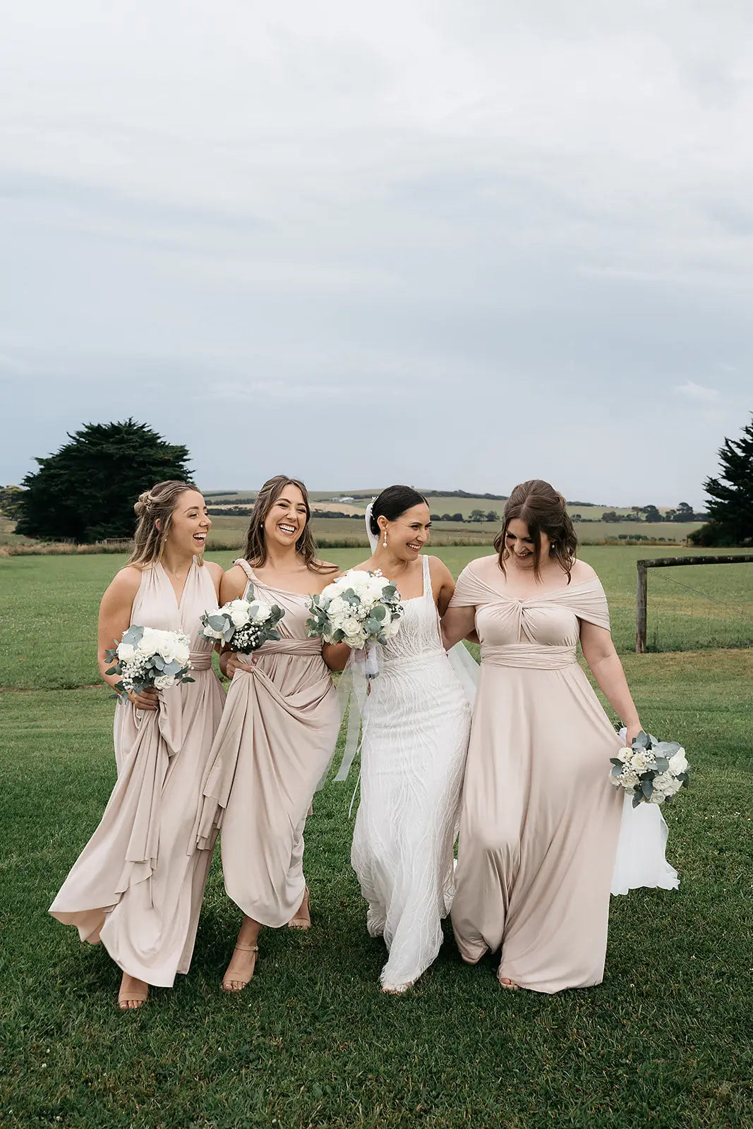 Four women in wedding attire wearing infinity dresses walking across a grassy field
