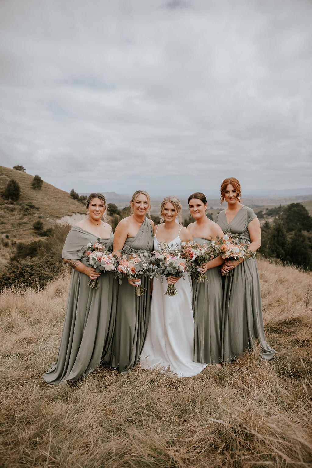 Bride in a white gown with four bridesmaids in matching Sage Infinity Gown multiway bridesmaid dresses holding floral bouquets