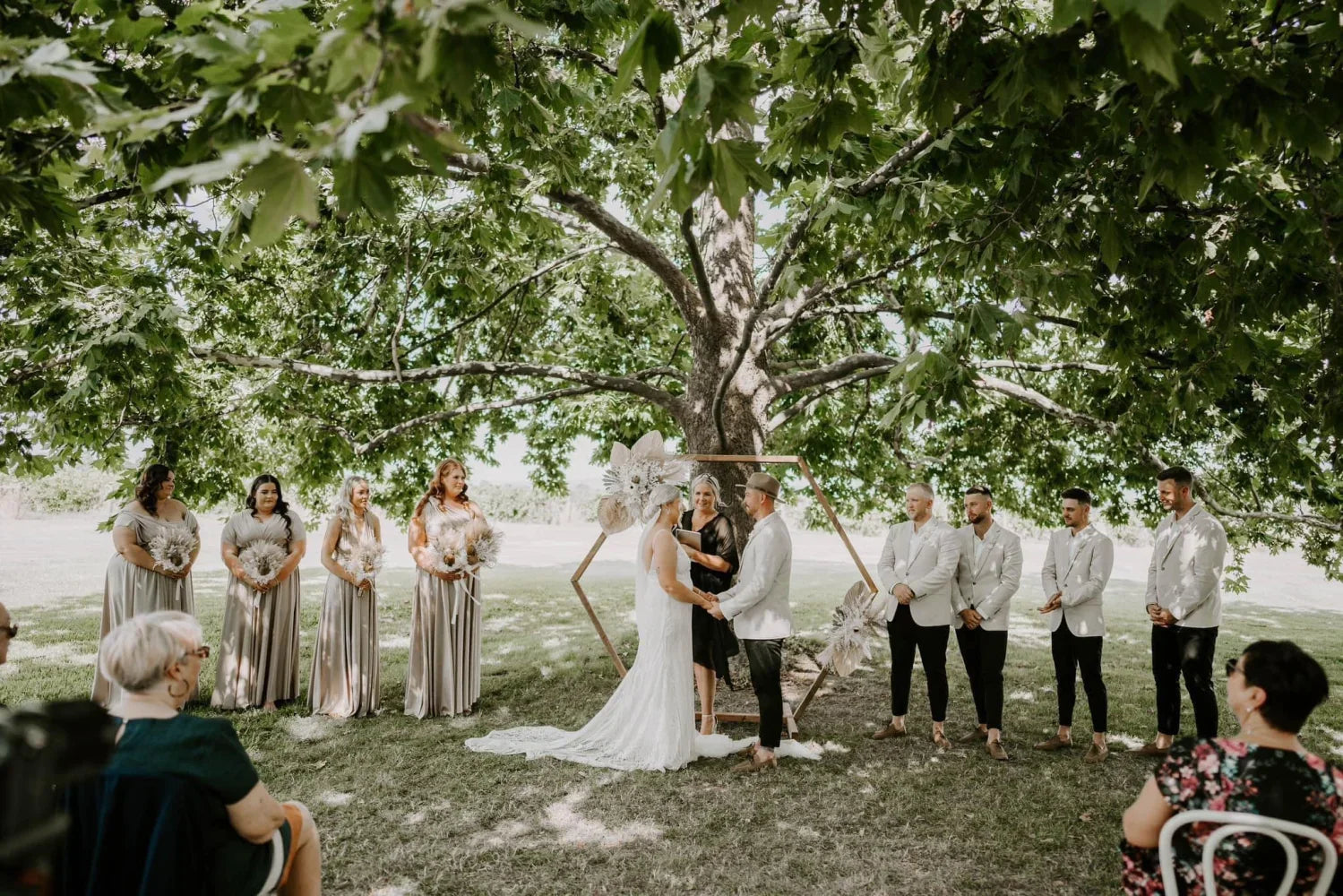 Outdoor wedding ceremony under a large tree featuring a Gold Infinity Gown, perfect bridesmaid dress Australia