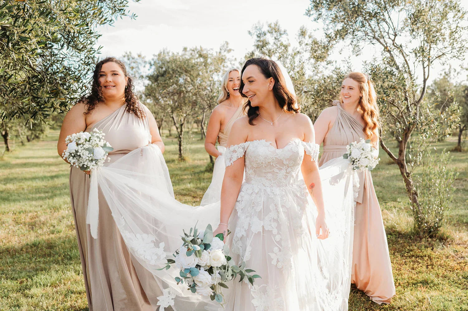 Bride in white lace infinity gown with bridesmaids in beige and pink dresses outdoors
